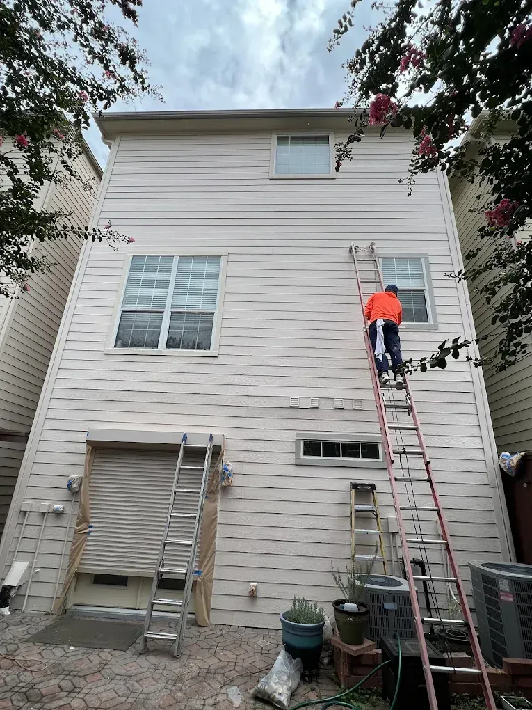 A person on a tall ladder working on the exterior of a two-story beige house. 