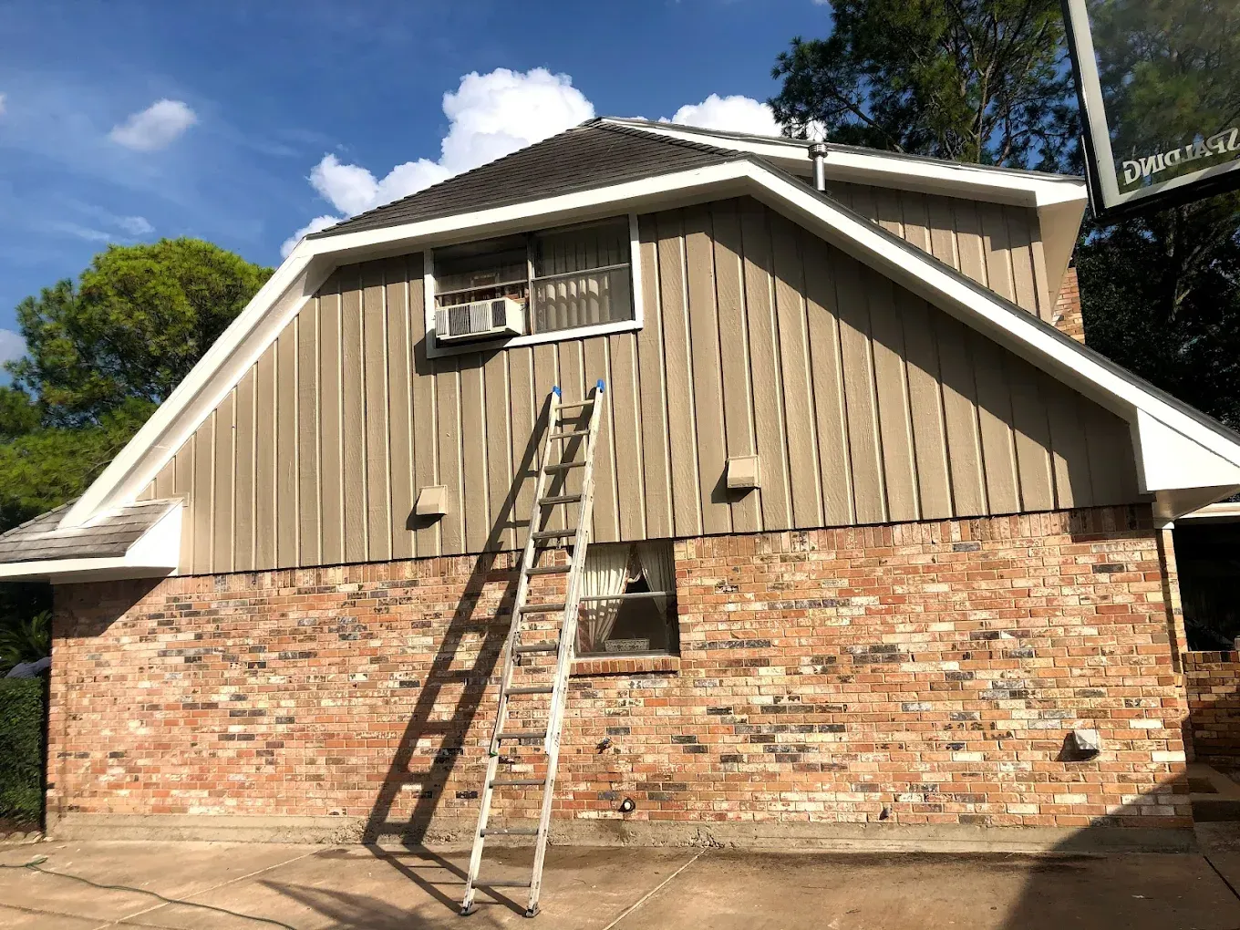 A house with a brick base and tan siding. A ladder leans against the side. The sky is blue.