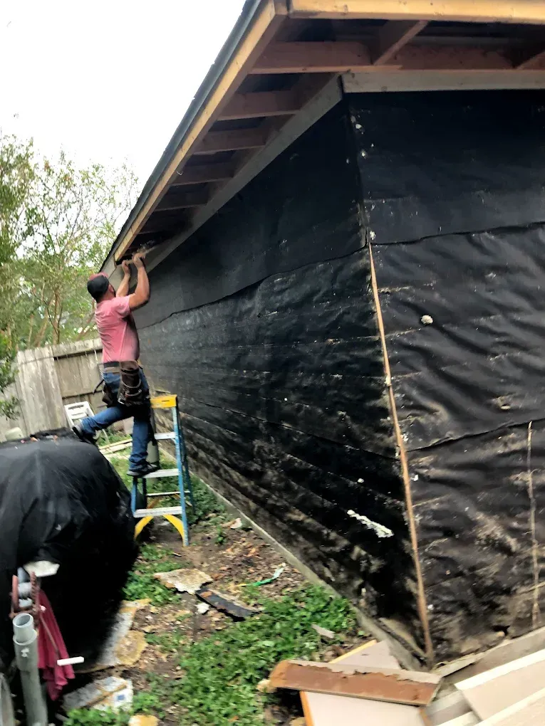 A construction worker on a ladder attaching something to the side of a building covered in black material.