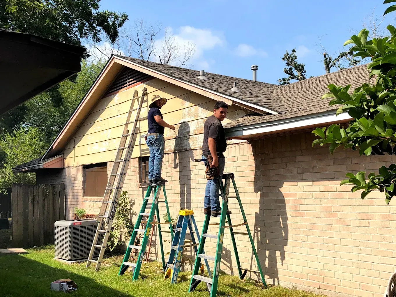 Two workers on ladders repair the siding of a house. The house is tan with a brown roof, under a bright blue sky.