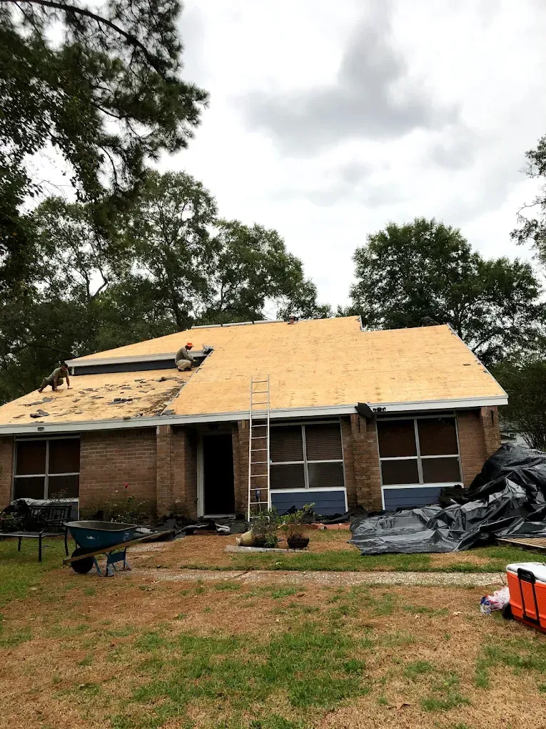 House with partially replaced roof; workers on the roof, exposed wooden boards visible. Dark brick exterior; cloudy sky.