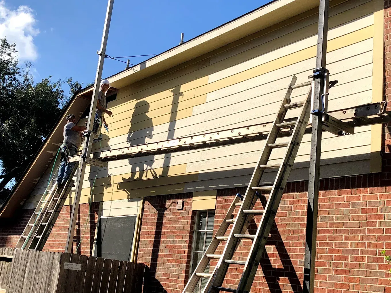 Two workers on ladders repair light yellow siding on a brick building. The setting is outdoors, sunny.
