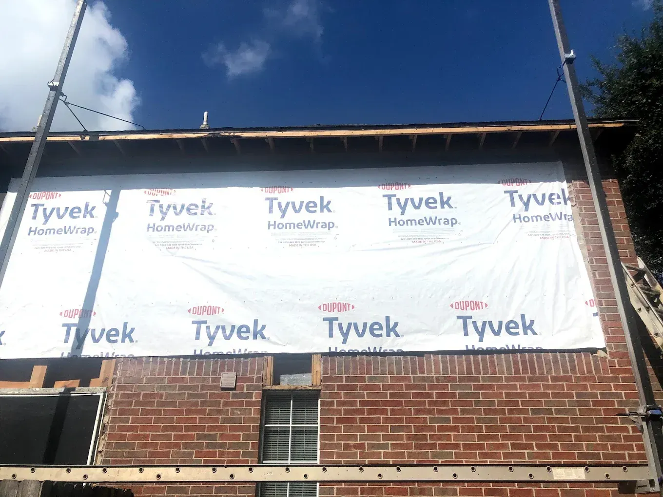 Exterior brick wall with Tyvek house wrap, scaffolding, and clear blue sky. Construction or renovation in progress.