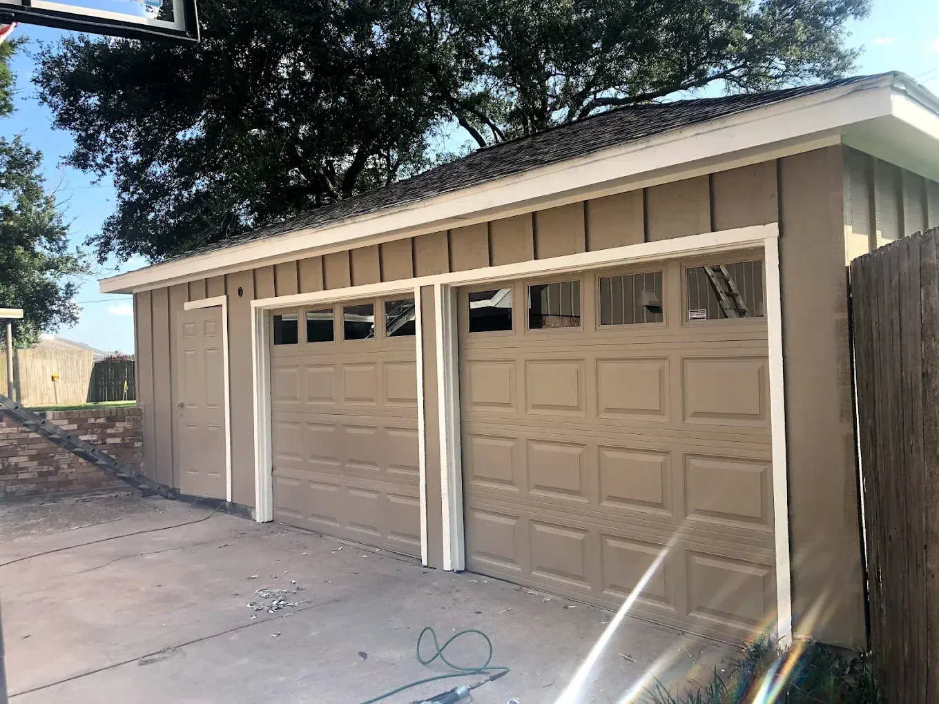 Tan garage with two doors and a side door. The trim is white. A dark roof with foliage in the background.