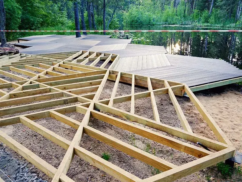 Wooden deck under construction next to a pond. Framing is visible, with some planks already laid. Trees in the background.
