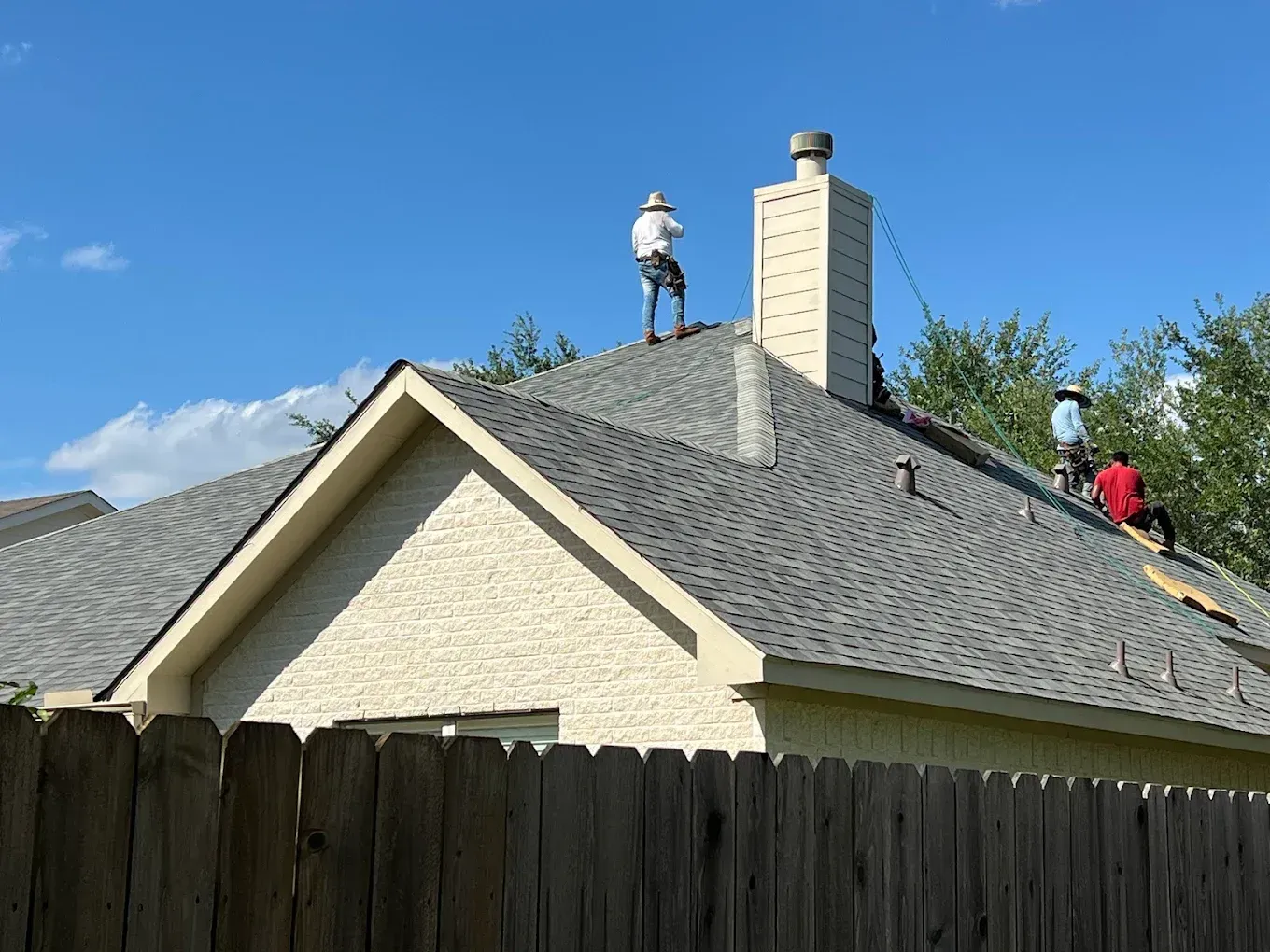 Roofers working on a gray shingled roof next to a tall white chimney on a sunny day. A wooden fence is in the foreground.
