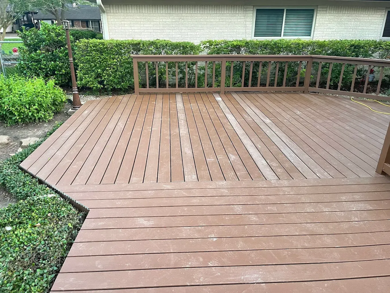 A brown wooden deck with warped boards and a railing, alongside green bushes and a house in the background.