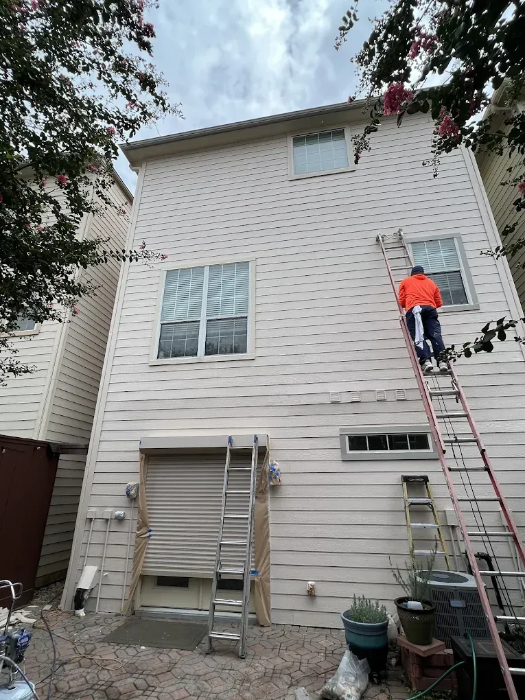 A person on a ladder painting the side of a two-story house. The siding is beige, and the sky is overcast.