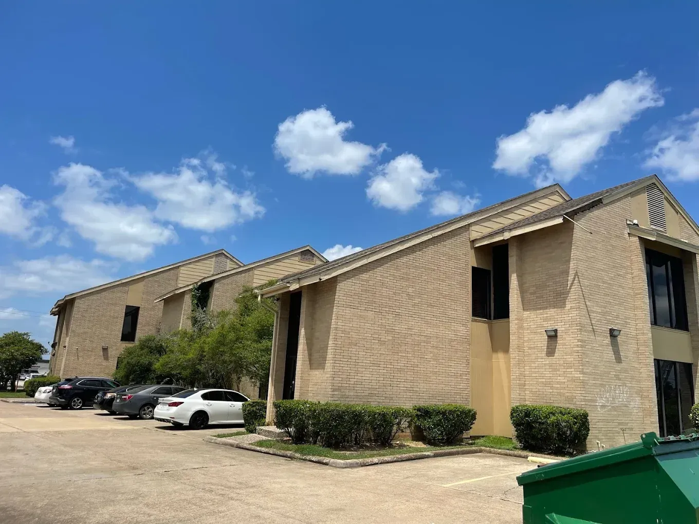Two-story beige apartment building with a parking lot under a partly cloudy blue sky. Several cars are parked in front.