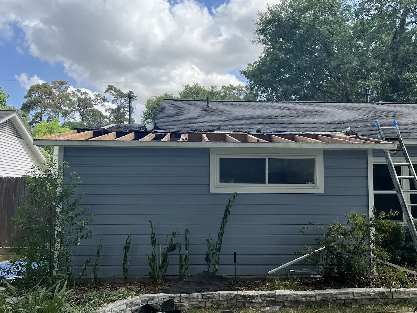 A house with a partially removed roof, showing exposed wood and the remaining dark shingles. A ladder leans against the side.
