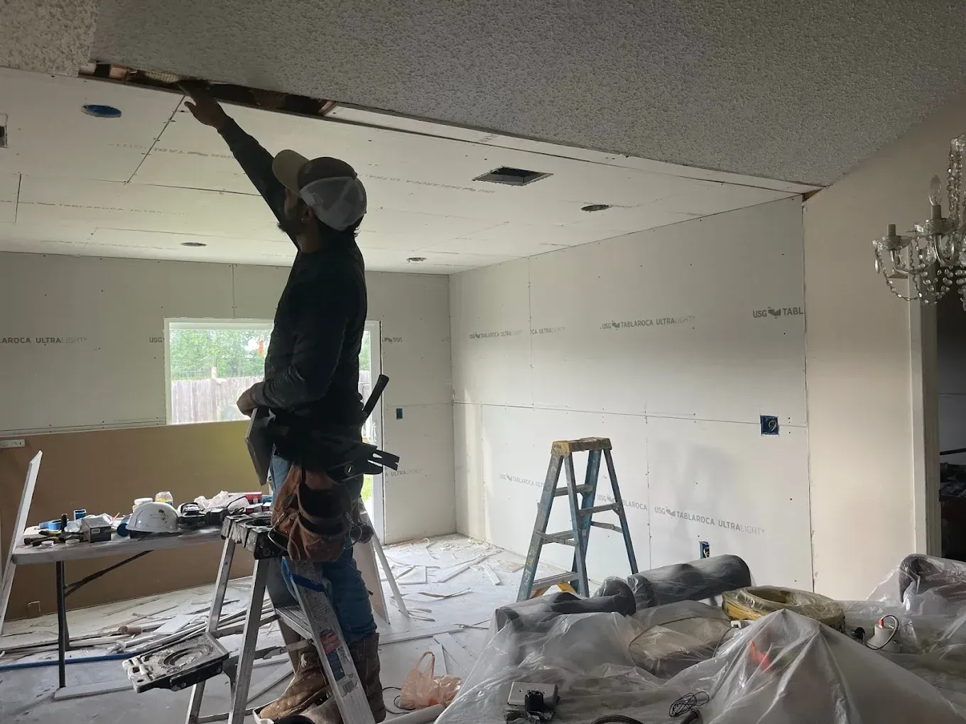 A person on a ladder installs drywall in a room with a new ceiling.