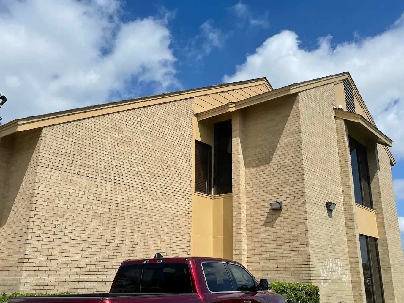 Tan brick building with a red truck parked in front, set against a blue sky with fluffy white clouds.