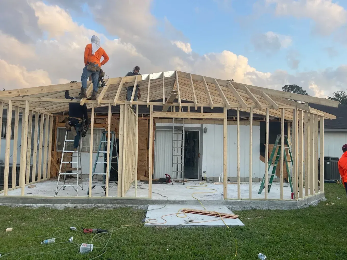 Construction workers framing an addition to a white house with a sliding glass door, under a cloudy sky.