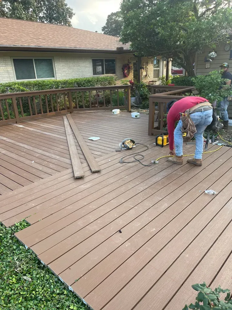 A carpenter works on a composite deck, using tools while bending over. The deck is brown, and a house is in the background.