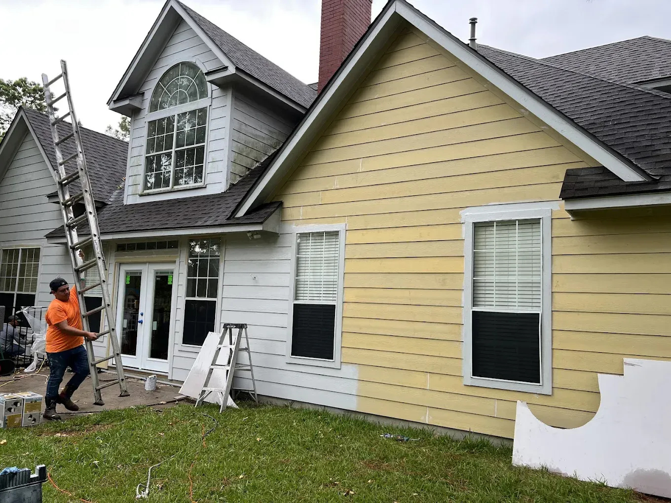 Man paints a two-story house, half painted yellow and the other half white. 