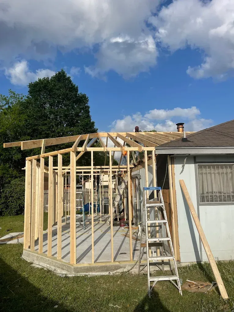 A wooden frame extension being built onto a house. The sky is blue with clouds, and the grass is green.