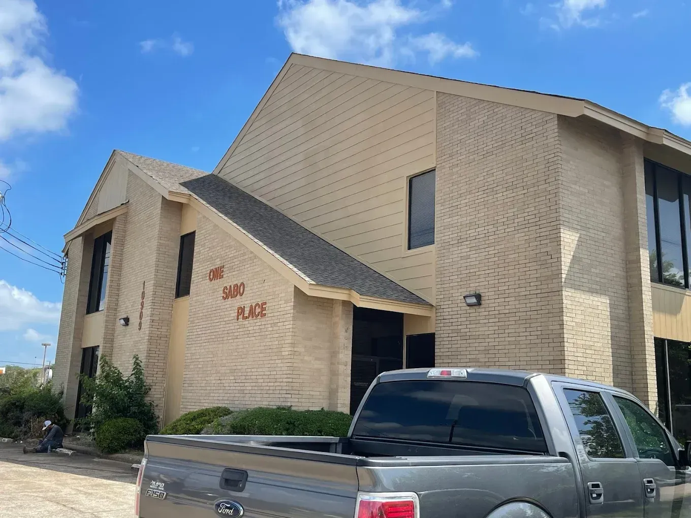 Beige brick building with dark roof, window, and truck parked in front.