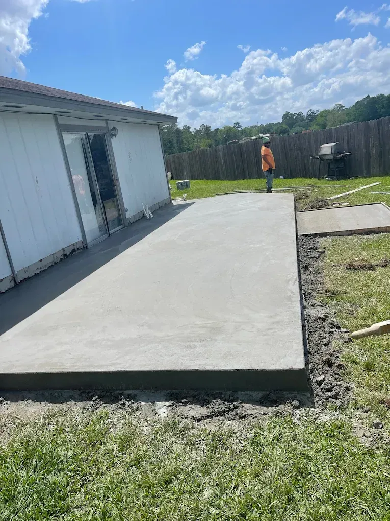 A newly poured concrete patio next to a white building, with a worker standing nearby in a grassy yard under a sunny sky.