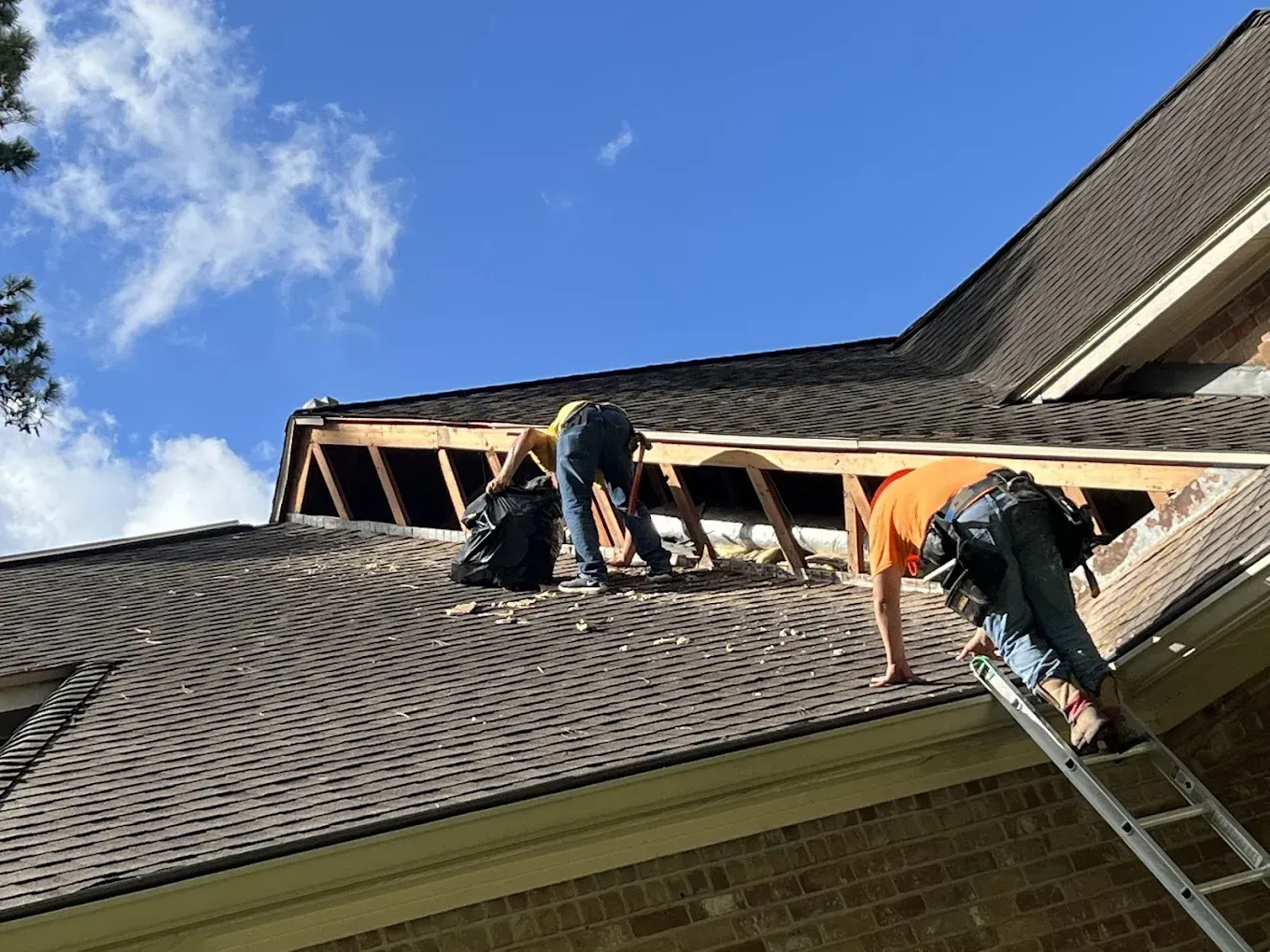 Two roofers on a roof removing shingles, bright sky overhead. One on ladder, the other leaning into the opening.