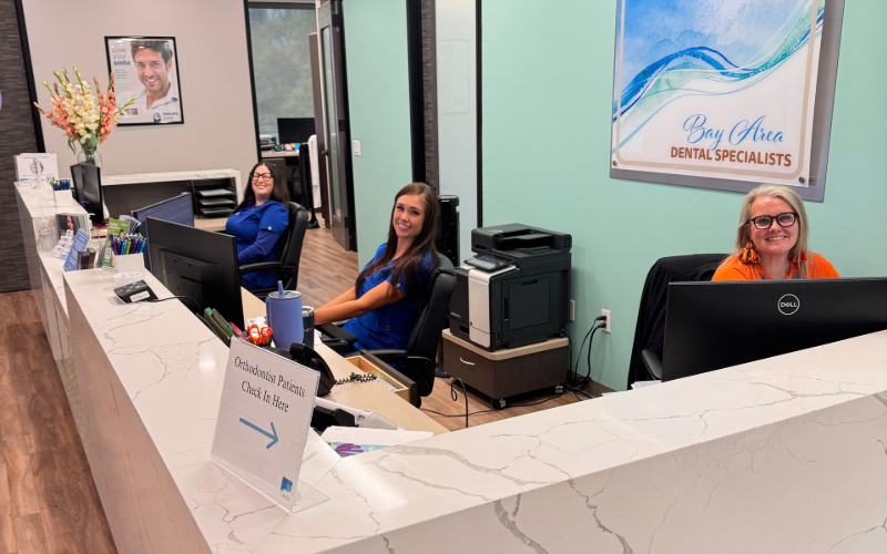Three staff members in professional attire smile from behind a long white marble reception desk in a brightly lit office.