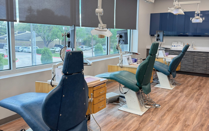 An open-concept dental office with three dark teal exam chairs facing a window with grey blinds and wood-look flooring.