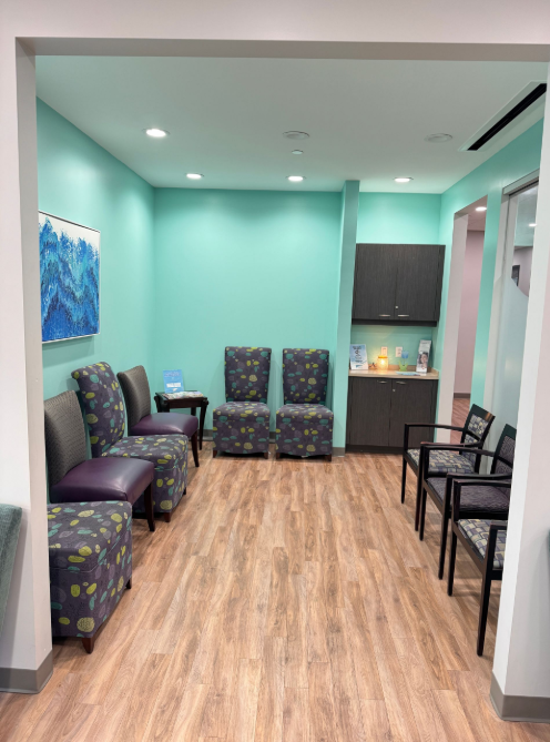 A waiting room with teal walls, patterned chairs, and a small refreshment area on the right.