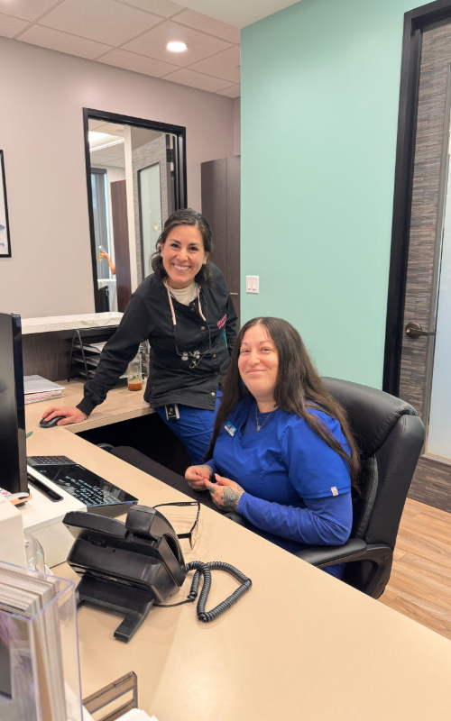 Two smiling office staff members in blue scrubs work behind a front desk in a bright, modern reception area.