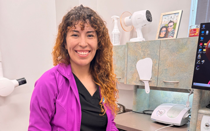 A smiling person with curly brown hair wearing a bright pink jacket in a dental office setting.