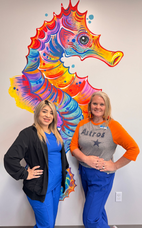 Two people in scrubs stand smiling in front of a wall featuring a large, colorful seahorse mural.