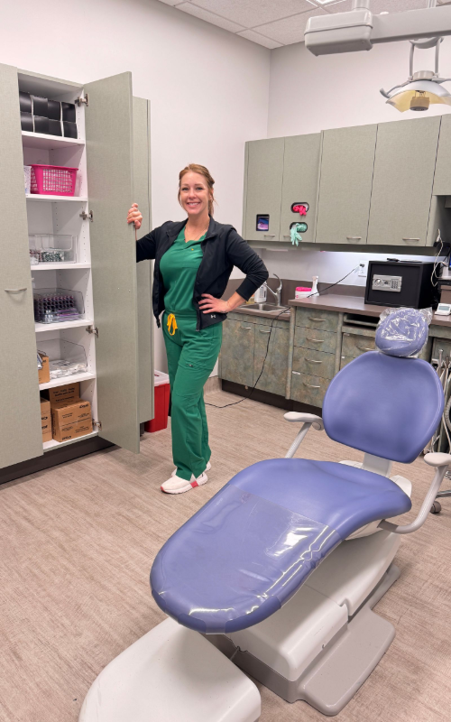 A person in green scrubs and a dark jacket stands smiling next to an open supply cabinet in a dental office.