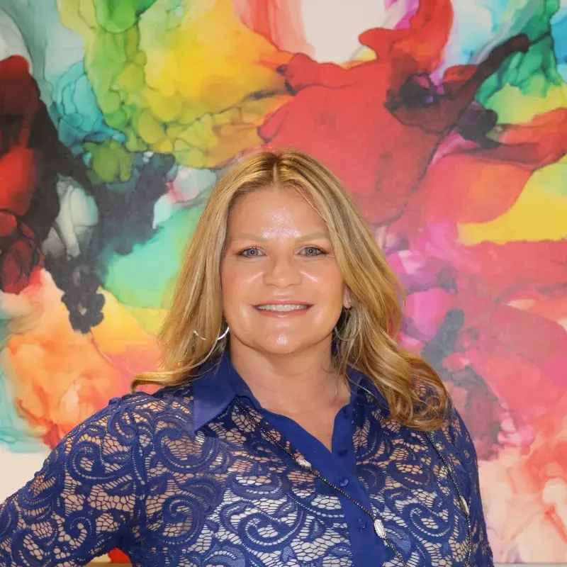 A woman in a blue lace shirt is smiling in front of a colorful painting