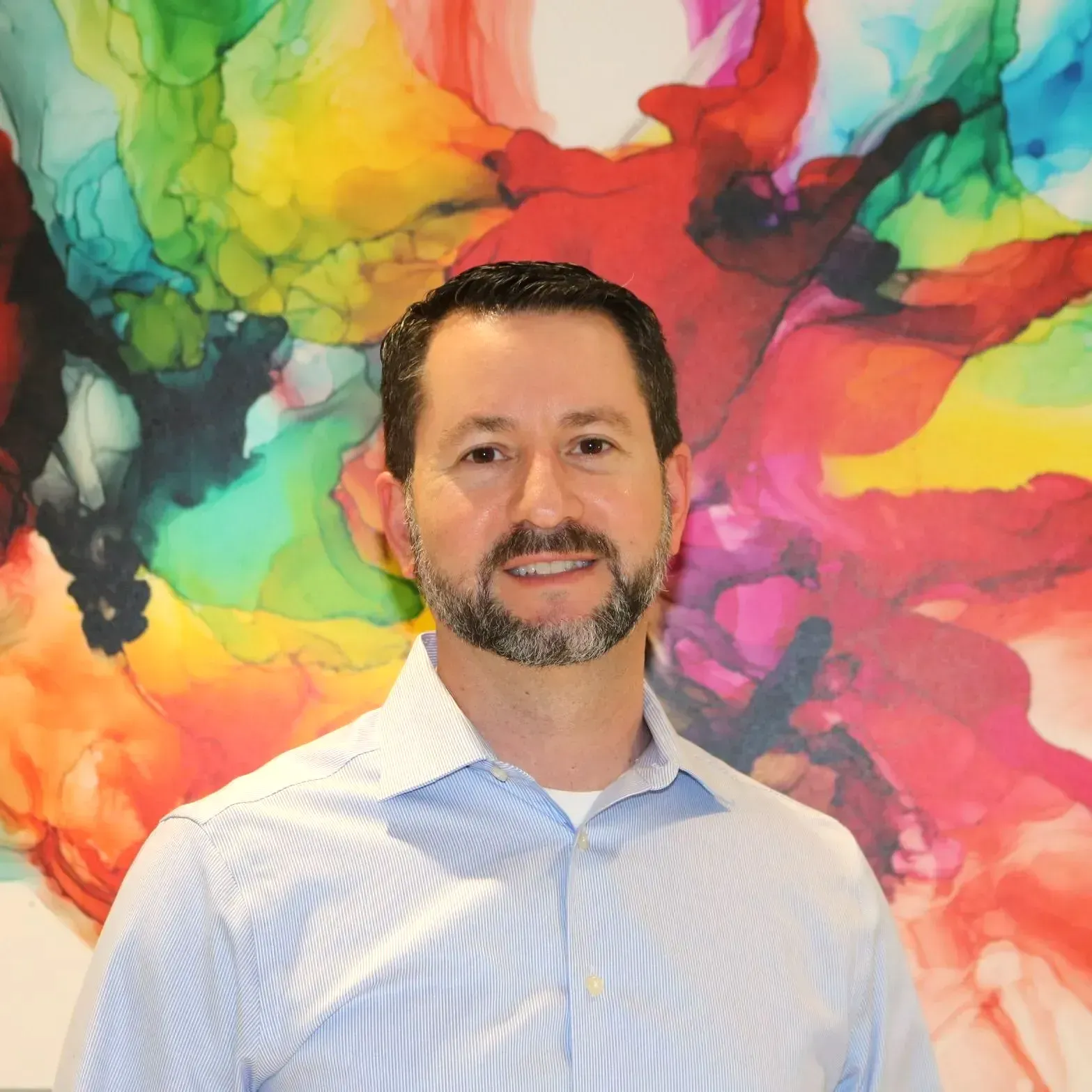A man with a beard is standing in front of a colorful painting.