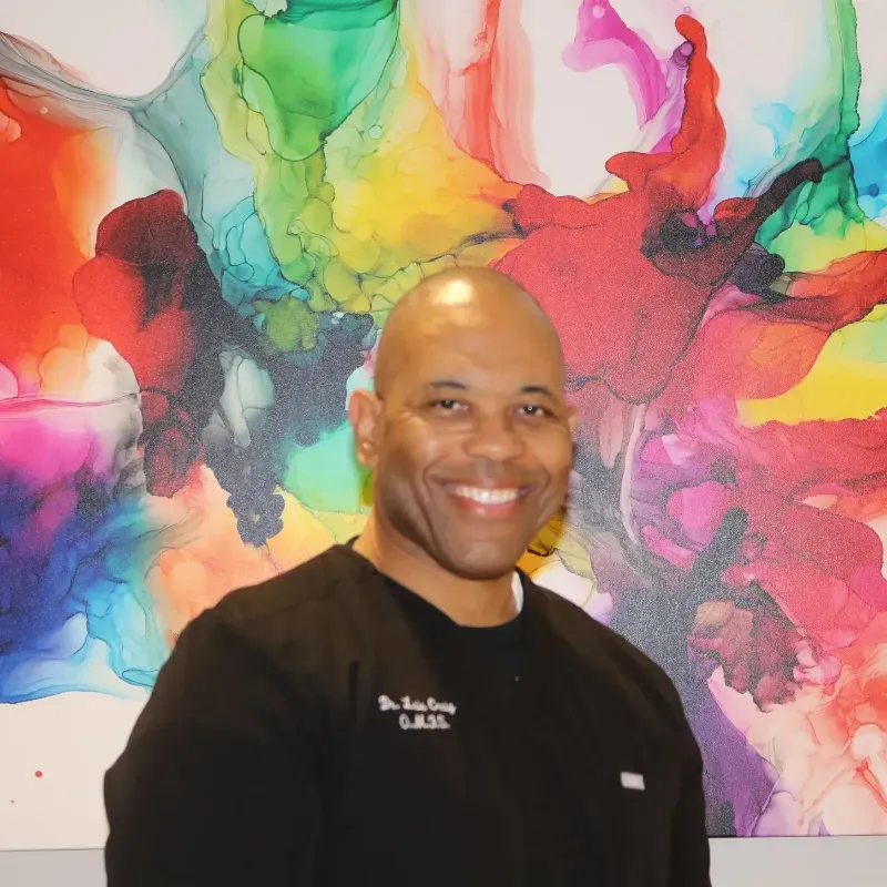 A man in a black shirt with the name g.a.b.s. on it smiles in front of a colorful painting