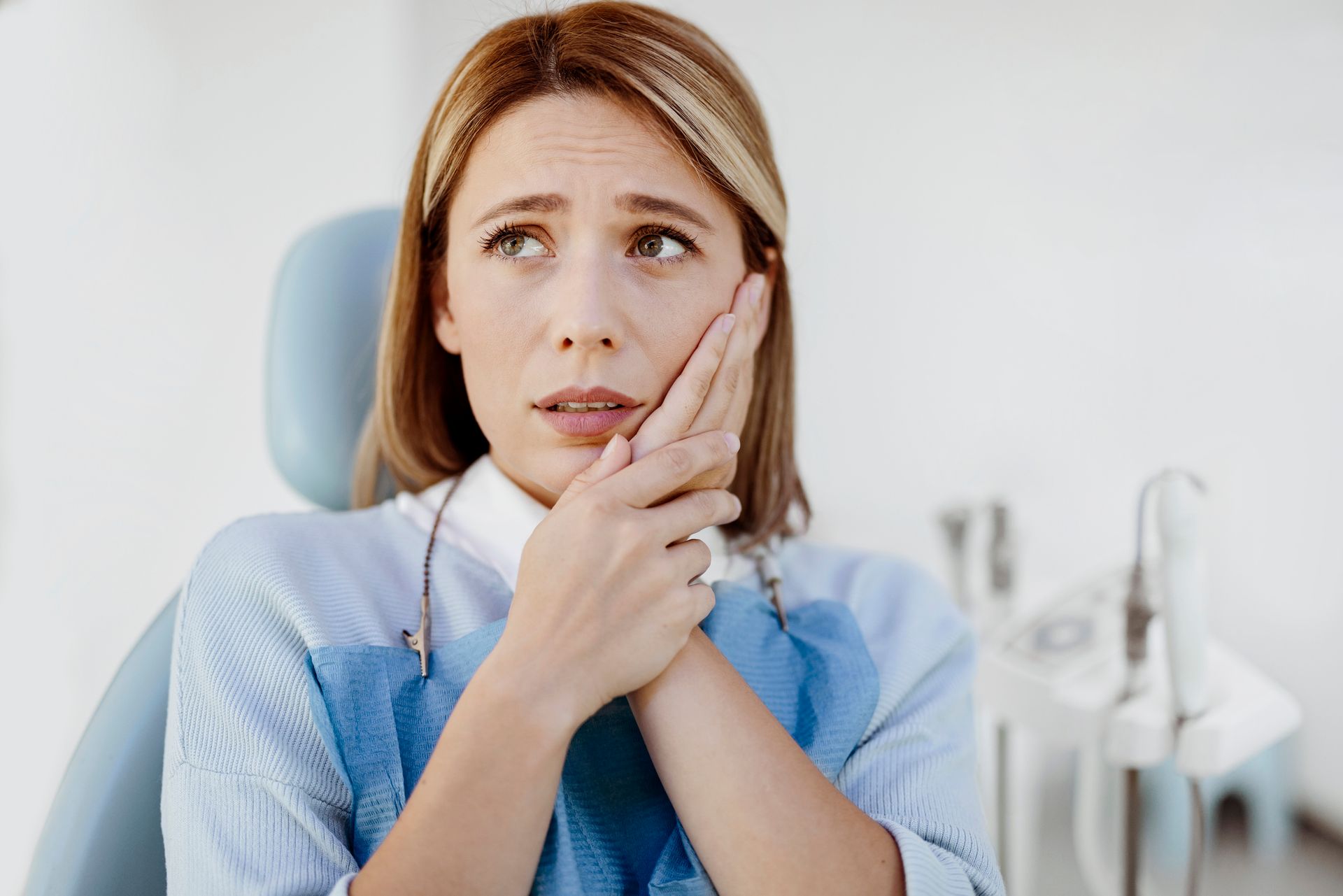 Woman in dental chair holding her jaw, looking pained. Light blue dental bib, white background.