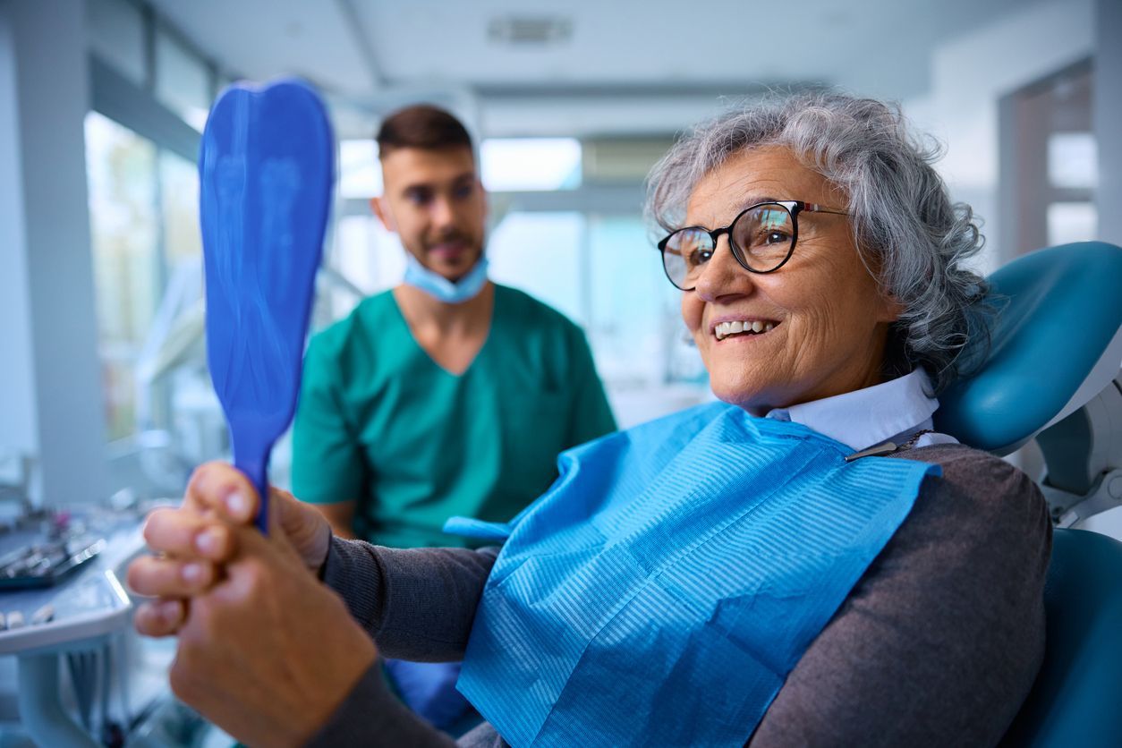 Woman in dental chair smiles while looking in a hand mirror; dentist in background.