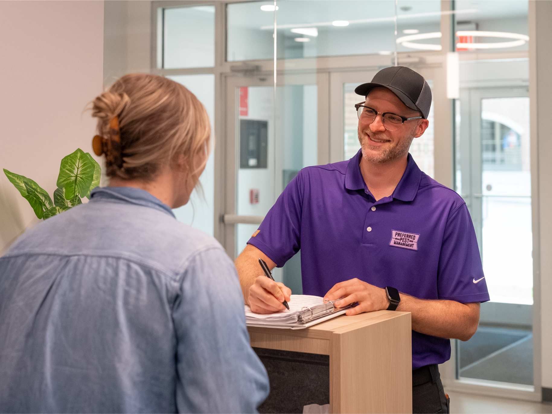 Man in purple shirt smiles at woman at a reception desk, taking notes.