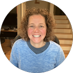 Woman with curly brown hair smiles, wearing a blue sweater. Interior setting with stairs visible.