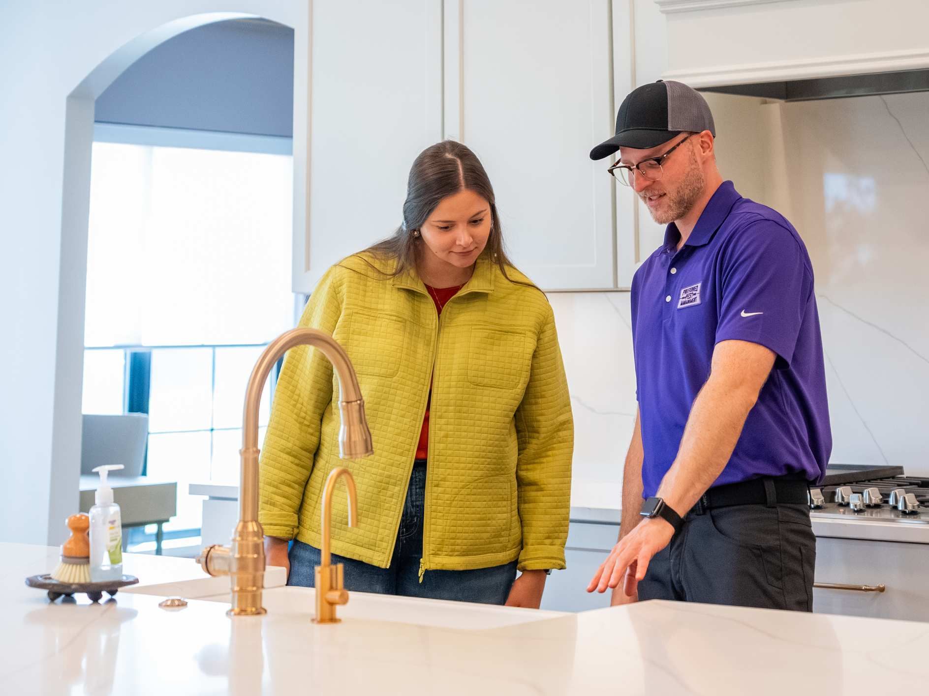 A woman and a man examine a kitchen faucet. The man points at a counter.