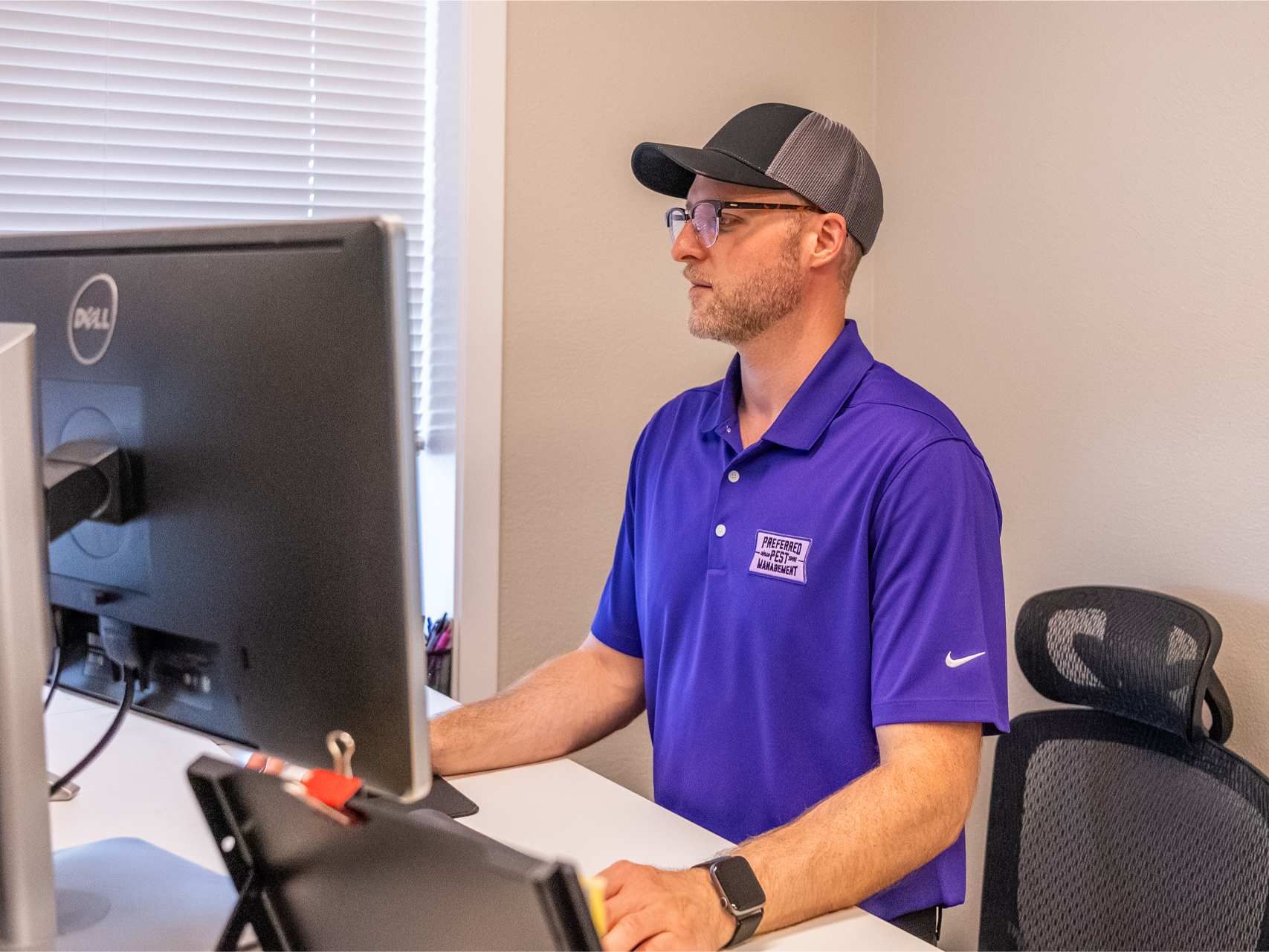 Man in purple shirt, cap, and glasses working at computer in office.