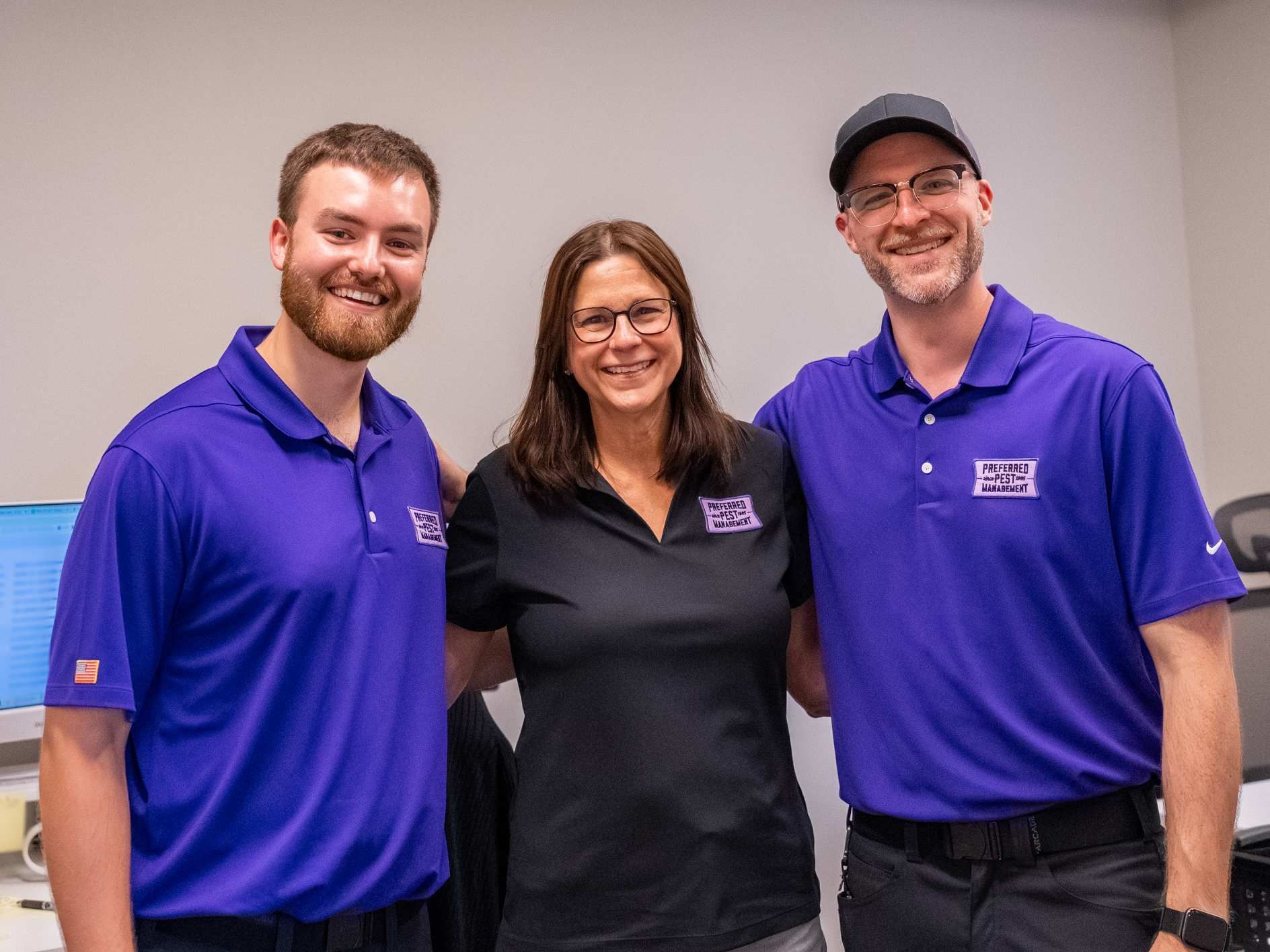 Three smiling people in a room: two men in purple shirts and a woman in a black shirt, arms around each other.