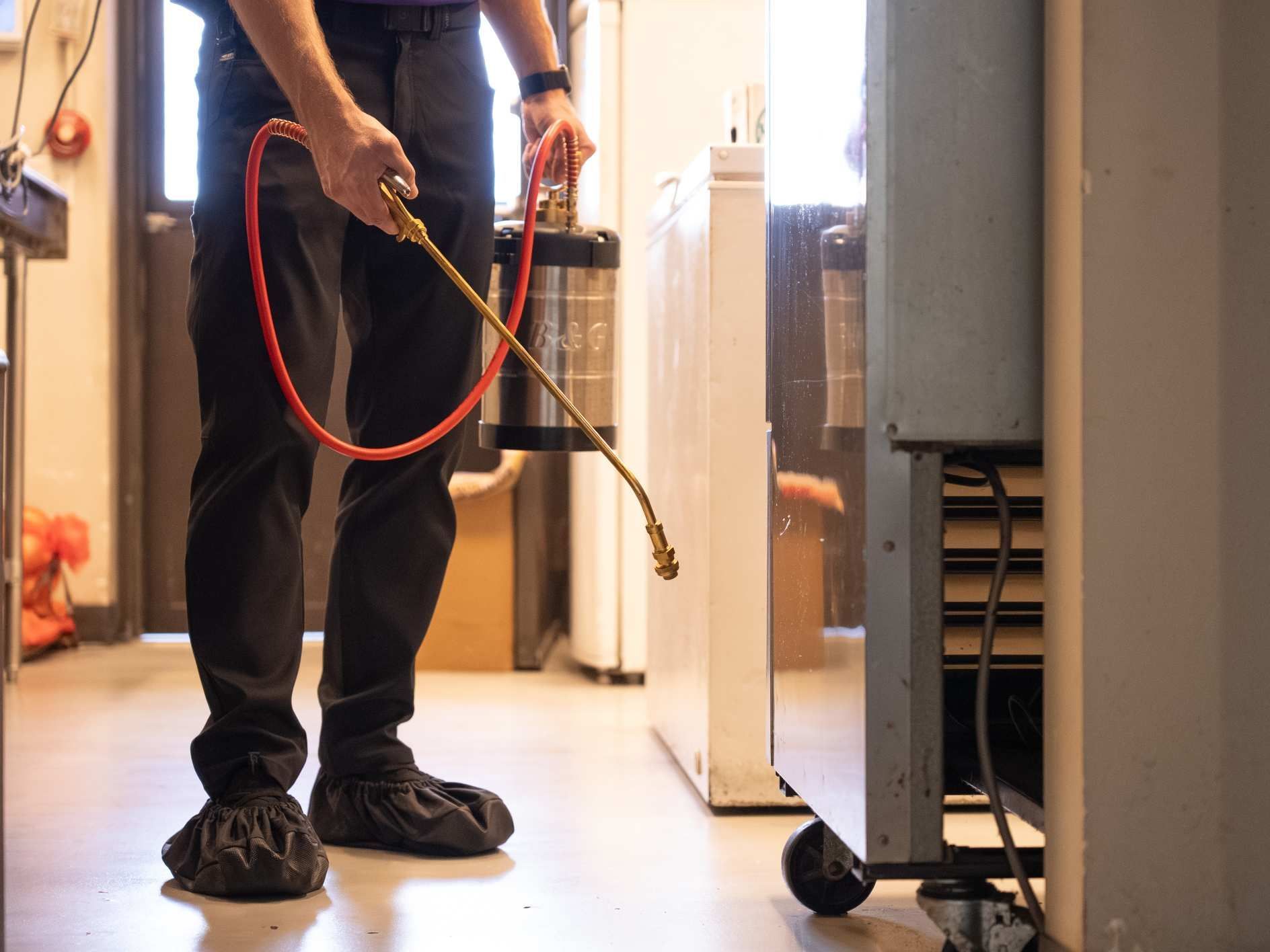 Person spraying insecticide near a refrigerator, wearing shoe covers indoors.