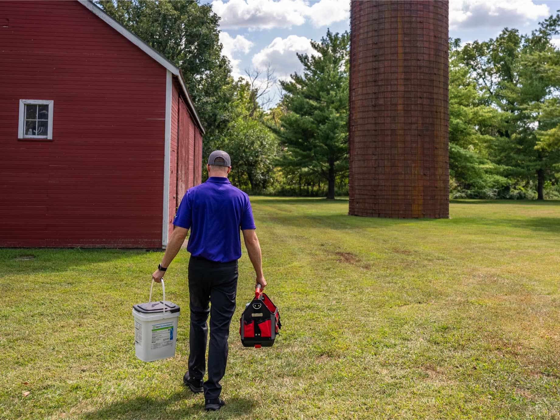 Man walking on grass towards a red barn and a tall brown tower, carrying a bucket and toolbox.