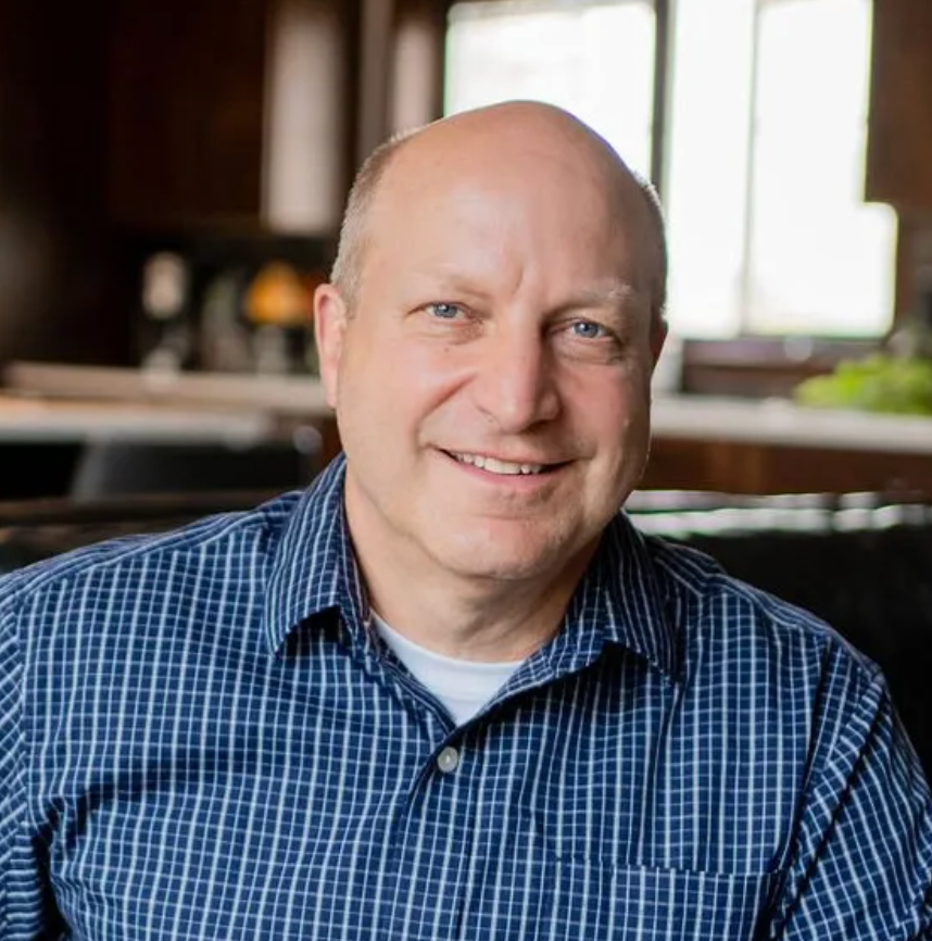 Bald man with blue checkered shirt smiles indoors.