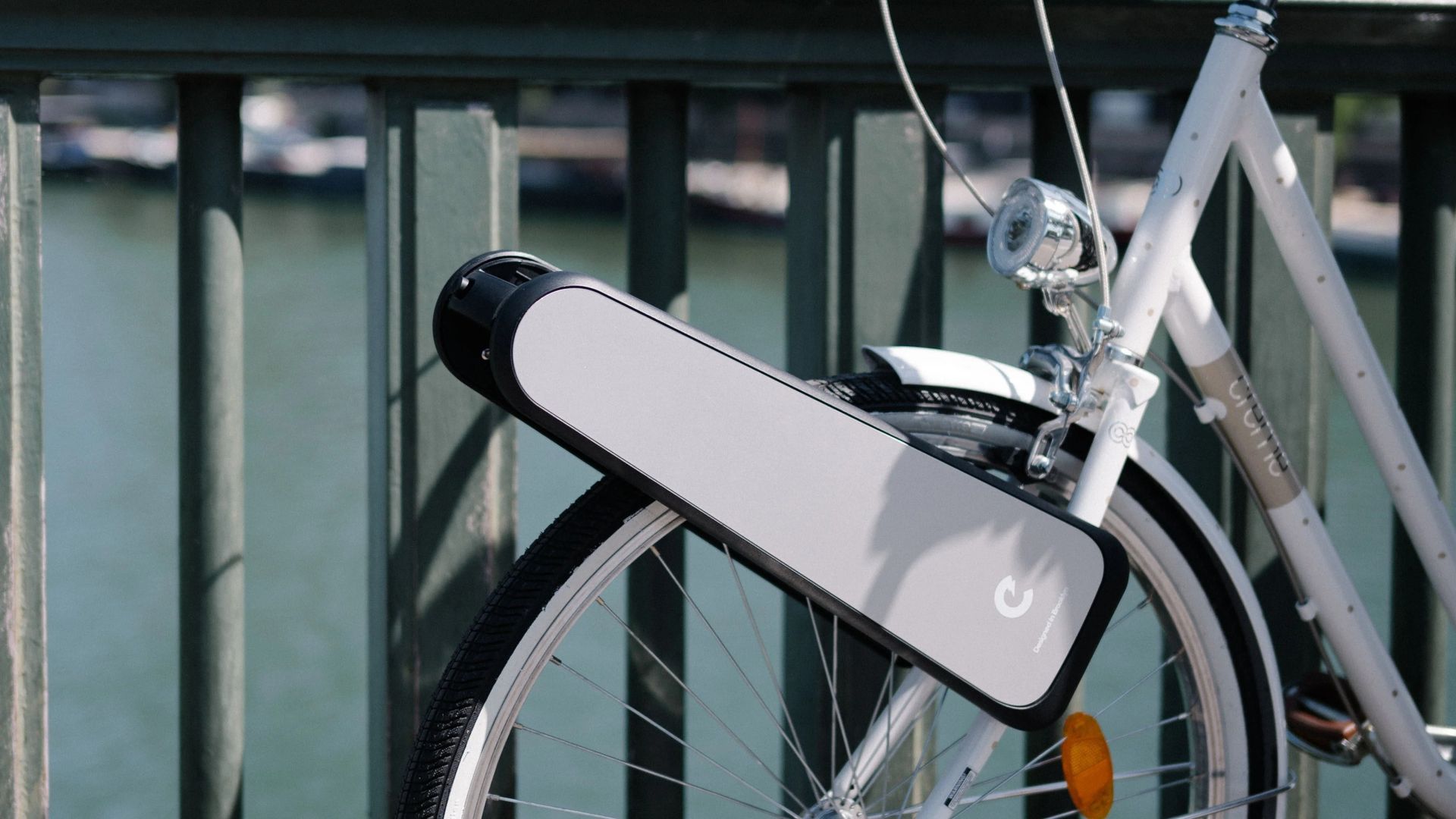 White bicycle with a large, rectangular device mounted on the rear wheel, near a metal railing.