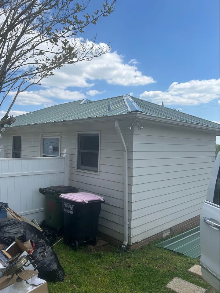 A small white house with a green roof and trash cans in the backyard.