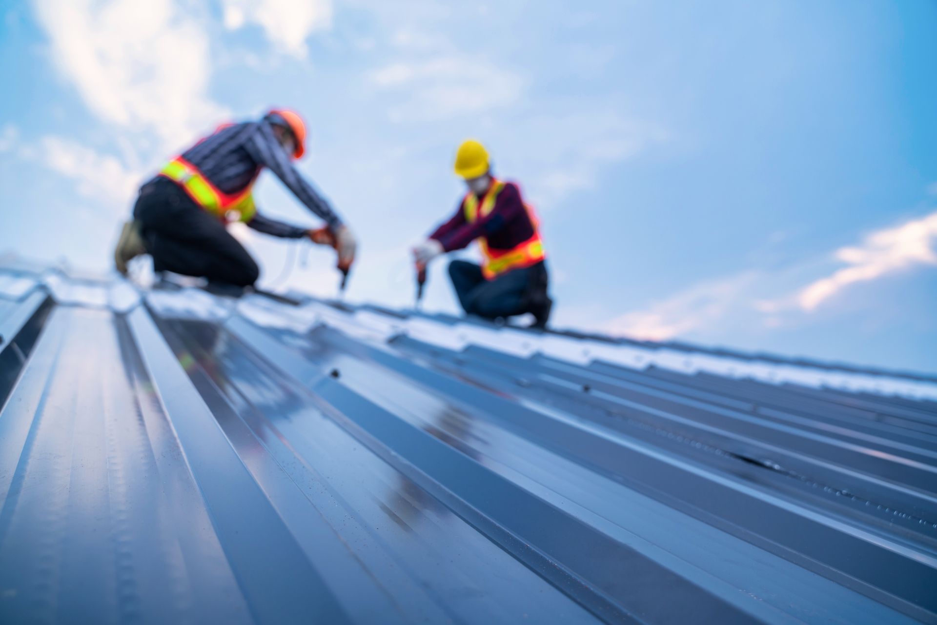 Two workers in safety gear installing metal roofing on a steep roof under a blue sky