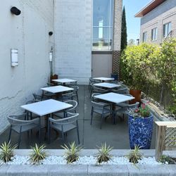 Outdoor café patio with white square tables, gray chairs, and potted plants beside a building and hedge