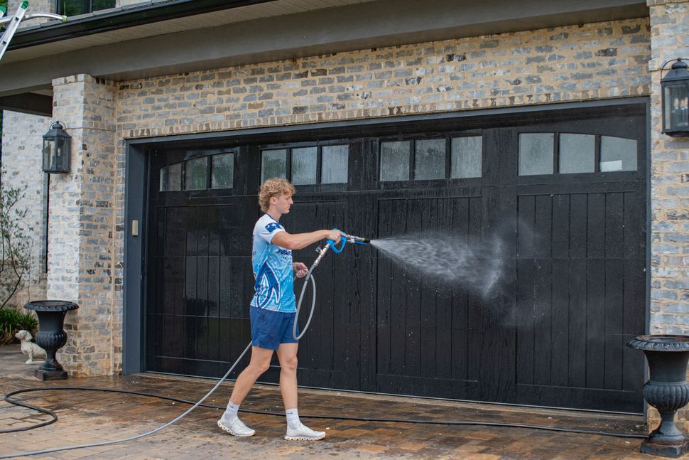 Person pressure-washing a black garage door in front of a brick house