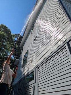 Person on a ladder cleaning a white house exterior with a long brush under a blue sky
