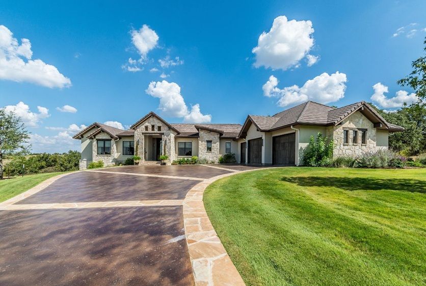 Large stone house with long driveway on a sunny day. Green lawn, blue sky with clouds.
